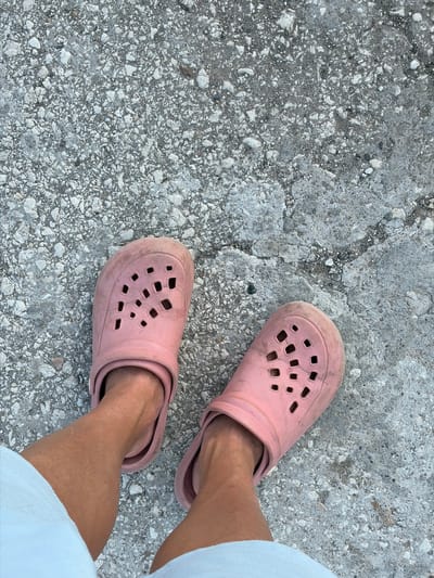 A pair of dirty, pink crocs are worn by an individual standing on a rough, grey concrete surface with visible gravel and cracks. The viewpoint is from directly above, focusing on the feet and lower legs, which are partially obscured by light-colored clothing. The context of Dolni Dabnik, Bulgaria is not directly discernible from the visual information in the image.