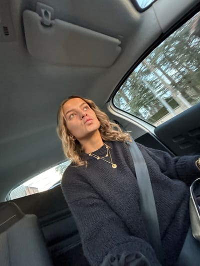 A woman sits in a car, wearing a black sweater and a constellation necklace. She has blonde hair and is looking at the camera with a slight pout. She is wearing a seatbelt. The car's interior is visible, including the sun visor and the headrest. Outside, trees and buildings are seen through the window. The photo is taken from a slightly angled perspective.