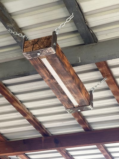 A homemade wooden object, possibly a light fixture or shelf, is suspended by chains from the underside of a metal roof structure supported by wooden beams. The wooden object is rectangular and appears to be constructed from stained and weathered wood. It has metal brackets at each end where the chains attach. The roof is made of corrugated metal sheeting, and the beams are rough-hewn timber, some appearing dark and aged. The scene is outdoors, likely under a covered porch or carport, given the visible roof and supporting structure. There are no people or animals present in the image. The location of Tabuny, Russia is not visually discernible from the provided image.