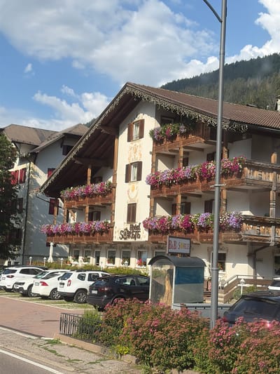 The image shows a hotel and B&B named "B&B SOREIE" in Pozza di Fassa, Italy. The hotel is a multi-story building with traditional alpine architecture, featuring wooden balconies adorned with numerous colorful flowers. The building has a brown tiled roof with decorative trim. A parking lot is visible in front of the hotel, with several cars parked, including white SUVs, a white sedan, and a black SUV. A lamppost stands in front of the hotel. The background shows a dense forest covering a mountain slope under a blue sky with white clouds. The scene appears to be captured during daytime.