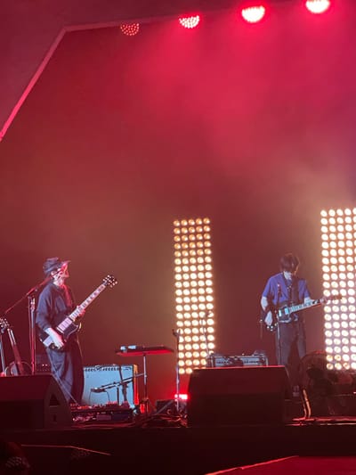 The image captures a music performance in Salembaran, Indonesia. Two guitarists are visible on stage, illuminated by a combination of red and white stage lights. One guitarist is wearing a blue jacket and playing a light-colored electric guitar, while the other is dressed in black and playing a dark electric guitar. Stage equipment, including amplifiers and a fan, is visible behind the musicians. The overall environment suggests a live concert setting.