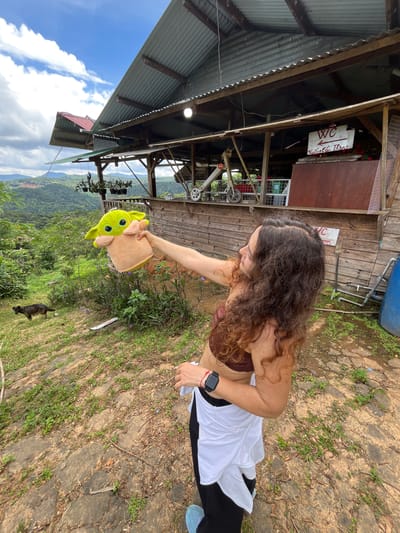 A woman with curly brown hair, wearing a brown crop top and black pants, holds up a plush toy of Grogu from Star Wars. She is standing outdoors on a rocky, grassy slope, with a wooden structure in the background. The structure appears to be a covered outdoor area with a corrugated metal roof and wooden walls. A sign with "WC" and Vietnamese text is visible on the wall. Beyond the structure, a lush green, mountainous landscape stretches into the distance under a cloudy sky. A dark-colored dog is visible in the background, to the left of the woman. There is also a blue plastic water barrel and some plumbing pipes to the right of the structure.