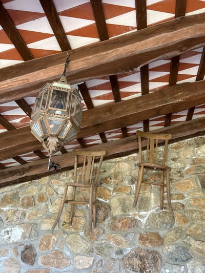 This image depicts an interior view looking upwards at a beamed ceiling and a stone wall. The ceiling is constructed of exposed wooden beams supporting a decorative pattern of alternating red and white triangular tiles. Hanging from one of the beams is an ornate, multi-faceted lantern made of glass and metal.
Attached to the stone wall are two simple wooden chairs, oriented sideways and appearing to be hung as decorative elements rather than for seating. The stone wall itself is composed of irregularly shaped stones of various earthy tones, cemented together. A small, dark metal decorative bird is attached to one of the wooden beams, near the lantern.
The scene suggests a rustic or traditional architectural style, possibly a patio, covered walkway, or interior space in a building with a stone facade. The lighting appears natural, indicating it's daytime. There are no people visible, and the overall mood is static and decorative. The provided location context places this scene in Tordera, Spain. No visible text is present in the image.