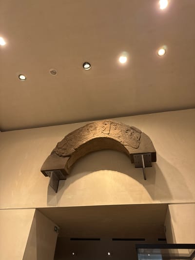 An ancient stone archway, carved with relief figures, is mounted on a cream-colored wall in a museum setting in the City of Edinburgh, United Kingdom. The arch is supported by two dark grey metal brackets attached to the wall. The ceiling above the arch features several recessed spotlights and one surface-mounted light fixture. The arch itself has a rough, weathered texture and displays faces with distinct features, as well as other carved designs that are partially obscured. Below the arch, a doorway or entrance is visible, with a cream-colored wall leading into a darker interior space. There are two green and white directional exit signs on the wall to the left of the opening. The overall lighting is dim, with spotlights creating distinct pools of light and shadow.