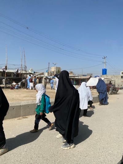 A street scene shows several individuals walking. A person in a long black robe leads, followed by a person in a patterned white shirt and a white head covering. A smaller child wearing a white head covering and carrying blue and green bundles trails behind. Electrical wires stretch across a blue sky. Buildings and parked motorcycles line the side of the road where people are gathered.
