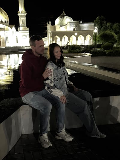 A man and a woman are sitting on a ledge at night in front of the White Mosque in Bolgar, Russia. The man is wearing a maroon hoodie and light blue jeans, with his arm around the woman. The woman is wearing a denim jacket over a white shirt and light blue jeans. They are both wearing athletic shoes. The White Mosque is illuminated and reflected in the water in front of them. Trees and lights are visible in the background. The overall impression is of a romantic setting at night.