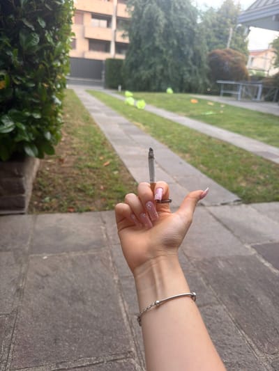A person is holding a lit cigarette in their hand. They have manicured nails with a floral design and are wearing a silver bracelet. The setting appears to be outdoors, next to a grassy area and a stone walkway. In the background, there are buildings, trees, and other vegetation. Given the context of Lissone, Italy, the location could be a residential or commercial area with green spaces.