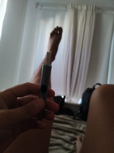 The image captures a close-up of a person's hand holding a grey electronic smoking device, possibly a heated tobacco product. The device has a black circular button on its side and a white filter-like component inserted into its top. In the background, a person's bare feet are visible, resting on a bed with striped bedding. Light-colored curtains hang behind, suggesting an indoor setting, perhaps a bedroom. The overall lighting implies it might be daytime, as natural light filters through the curtains. There are no visible signs of weather conditions. The scene appears relaxed and casual, possibly indicating a moment of leisure. The specific location of Germasogeia Municipality, Cyprus, is provided as context but is not visually evident in the image itself. There is no discernible text in the image.