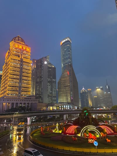 The image captures a twilight scene in Pudong, China, likely during or after a rain shower, given the wet roads and reflections. The skyline is dominated by towering skyscrapers, many of which are illuminated.
On the left, a prominent building with a classical dome is lit in warm yellow and features bright red Chinese characters on its facade that read "中国平安" (China Ping An). To its right, a modern, slender skyscraper with a rounded top is also illuminated. Further to the right, other skyscrapers of varying heights and architectural styles fill the frame, some with their windows lit.
In the foreground, a circular roundabout is elaborately decorated with floral displays and illuminated sculptures. These decorations include arched structures outlined in red lights and several horse figures, some of which appear to be part of a display for the year 2024, indicated by a blue sign with the number "2024" and a circular arrow.
Below the skyscrapers, a multi-level road infrastructure is visible, with cars and a yellow double-decker bus navigating the wet asphalt. The reflections of the lights on the wet road create a dynamic and visually striking effect. The overall atmosphere is one of a bustling urban environment at dusk, with the city lights creating a vibrant spectacle against the darkening sky.