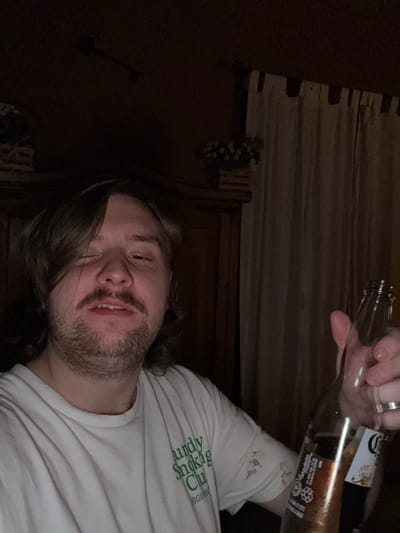 A person is holding two Corona beer bottles in a dimly lit room. The person has long, dark hair, a mustache, and a beard, and is wearing a white t-shirt with green text. The background appears to be a dark wooden wall, partially obscured by a patterned object. The location is unclear, but the image caption mentions Cañada de Madero, Mexico. The image is taken from a tilted angle.