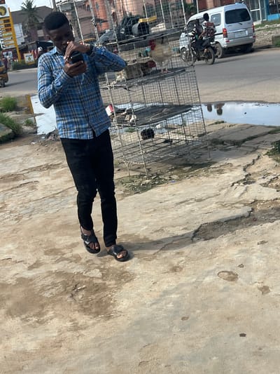 A man stands on a concrete surface in what appears to be an outdoor setting in Ijegun, Nigeria. He is wearing a blue plaid shirt, black pants, and black sandals. He is holding a mobile phone up to his face.

Behind him is a row of wire cages. Beyond the cages, there is a street with parked vehicles, including a white van and motorcycles. There are also buildings and trees visible in the background. The ground is cracked and discolored. A puddle of water is visible on the street.