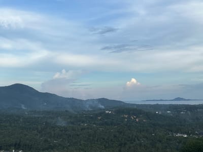 The image shows a landscape scene in Baan Hin Kong, Thailand, featuring a view from an elevated position. The foreground is dominated by dense, dark green vegetation, likely a forest or jungle. Smoke plumes rise in several locations amidst the trees, suggesting possible fires or controlled burns. A mountain range is visible in the midground, extending across the frame. In the background, there is a body of water, with a distant island. The sky is partly cloudy, with a mix of light blue and white clouds.