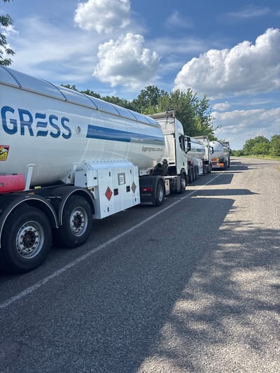 In Pereiaslavske, Ukraine, a line of white tanker trucks with blue trim are parked on the side of a paved road. The trucks have the word "GRESS" and "lpgprogress.com.ua" written on their sides. The sky is blue with white clouds, and trees line the side of the road. One truck in the foreground has a red container and warning labels on its side.
