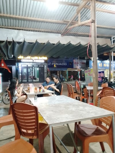 The image depicts an illuminated outdoor or semi-outdoor dining area at night in Thôn 8, Vietnam. The scene is illuminated by overhead fluorescent lights and features a corrugated metal roof supported by wooden beams. In the mid-ground, three individuals are seated around a square white-topped table with drinks and dishes present. From left to right: a female with long dark hair, wearing a white shirt and dark shorts, is actively engaged with a mobile phone; a male wearing a dark blue shirt and eyeglasses is looking downwards, possibly at a device or the table; and a female with dark hair tied back, wearing a black t-shirt, is also focused on a mobile phone. The table contains multiple dark beverage bottles, glasses, and food items. Surrounding them are several empty brown plastic chairs and other similar tables. In the background, a bustling street is visible with various illuminated storefronts displaying Vietnamese text, including "PINACO," "HỘI TÌNH," and "LÊ NGỌC" on business signs. A green, pleated fabric awning hangs above some of these establishments. Motorcycles are parked on the street. A red electric fan is positioned near an empty table to the right. The overall environment suggests a casual, open-air eatery alongside a commercial street.