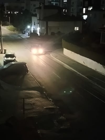 An elevated night-time view depicts an urban street scene in Nicosia, Northern Cyprus / Cyprus. The environment is illuminated by artificial light sources including streetlights, vehicle headlights, and interior lights emanating from structures. A two-lane asphalt road occupies the midground, featuring faded white lane markings and parallel lines consistent with tram tracks on its right side. A dark-colored vehicle with illuminated headlights is visible driving in the upper-middle section of the road, oriented towards the viewer. Two parked passenger vehicles are present on the left side of the road: a light-colored sedan and a partially obscured darker vehicle in the immediate foreground. A brick-paved sidewalk borders the left side of the road, hosting a street light pole and a light-colored refuse bin. On the right, a low white wall topped with a dense green hedge curves alongside the road. The background reveals multiple multi-story residential buildings, several with illuminated windows. A two-story house with a distinct rounded architectural feature is centrally located in the mid-distance. Two distinct, bright green reflective points are observed on the road surface in the lower-right quadrant.
