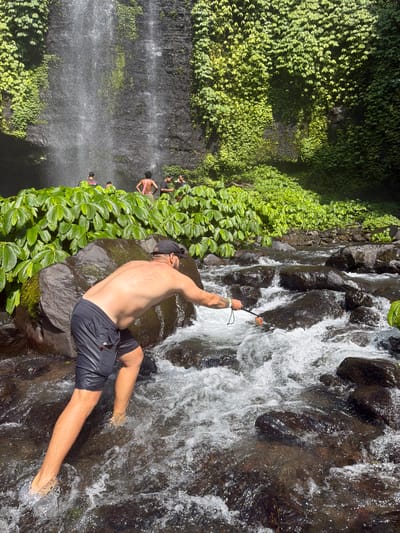 In the city of Sawan, Indonesia, at the base of a lush waterfall cascading down a dark, rocky cliff face, a man wearing black shorts and a black cap is navigating a shallow, rocky stream. He is bent over, with his right arm extended, holding a thin pole with a red object at the end. His body is positioned to his left, and his legs are submerged in the fast-flowing, clear water. Other individuals are visible in the background, standing near the waterfall and amidst dense green foliage that covers the rocky cliff. The stream bed is filled with dark, wet rocks of various sizes, and the water is turbulent, creating white foam. Sunlight illuminates the scene, highlighting the spray from the waterfall and the shimmering surface of the water.