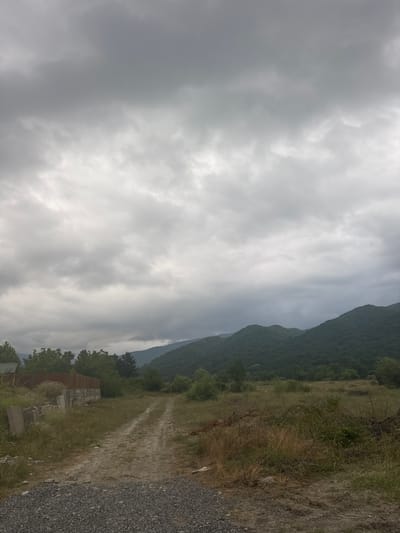 The image depicts a dirt road leading through a rural landscape in Aragvispiri, Georgia. The sky is overcast with heavy, gray clouds. In the foreground, the road is covered with gravel and dirt, with dry grass and some debris on the sides. To the left, there is a concrete fence and some scattered concrete blocks. Beyond the road, there is a field of tall, dry grass and scattered green bushes. In the background, rolling green hills covered with dense vegetation rise up, receding into the distance. The overall impression is one of a somber, perhaps damp, rural setting under a cloudy sky. There are no people or animals visible in the image.