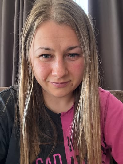 A medium shot photograph shows a young woman with long, blonde hair. She is wearing a split-colored shirt, with one half black and the other half bright pink. Her face is partially obscured by her hair. Her expression is ambiguous, with a slight frown and a hint of a smile. She has a small mole on her right cheek. The background appears to be a curtain or drapery, suggesting an indoor setting. No specific location information can be determined from the visual data.