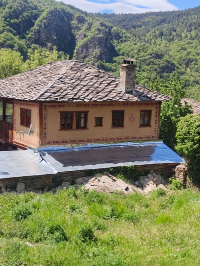 The image depicts a traditional house in Kovachevitsa, Bulgaria, situated amidst a lush, hilly landscape. The house features a stone roof, a stone chimney, and a beige facade with patterned trim around windows and along the top edge. There are several dark-framed windows on the side of the house facing the camera. The house is partially obscured by a metal roof structure on a lower level. A stone wall supports the structure. In the foreground, there is a field of green grass and various vegetation. Behind the house, a mountainside covered in dense green trees rises into the background. The sky is pale blue. The lighting suggests a bright, sunny day.