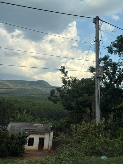 The image depicts a rural scene in Dak Ro Wa, Vietnam. An aged concrete utility pole with power lines stands prominently on the left. The sky is partly cloudy. In the background is a mountain shrouded in lush green vegetation. To the right, a small, weathered concrete building with a simple design sits partially obscured by foliage. The surrounding environment consists of thick vegetation characteristic of a tropical climate. No people are visible in the image.