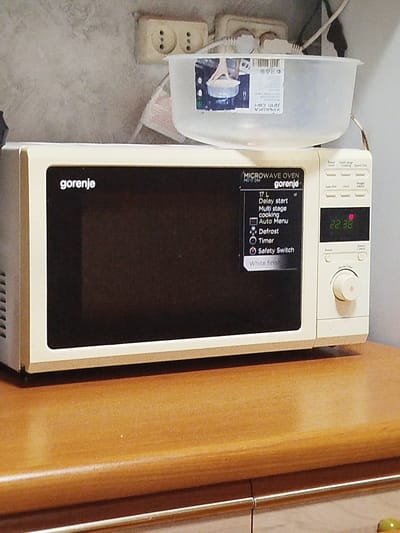 The image shows a Gorenje microwave oven, model MD 17 DW, with its control panel displaying "22:38". The microwave is positioned on a wooden surface, likely a countertop or shelf. Above the microwave, a white plastic container is visible, and behind it, an electrical outlet with two sockets is mounted on a textured wall. The overall environment appears to be indoors, possibly a kitchen. There are no people or actions depicted in the image. The city of Шой-Шудумарь, Russia is mentioned but not directly depicted or relevant to the visual content of the image.