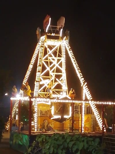 The image depicts a tall, brightly illuminated amusement park ride at night in Babban Giji, Nigeria. The central feature is a prominent lattice-work tower constructed from criss-crossing metal beams. Two large, arcing arms extend diagonally upwards and outwards from near the tower's apex, each terminating in a small, enclosed passenger pod; one pod appears reddish, the other lighter in color.

The entire ride structure, including the central tower, arcing arms, and a multi-level base platform, is extensively outlined with rows of glowing warm white string lights or LEDs, creating a striking illuminated pattern against the dark night sky.

On the upper level of the ride's lower platform, four static, humanoid figures are positioned. These figures appear to be decorative elements or mannequins rather than live individuals. Two figures are on the left, one standing and one crouching, wearing light-colored attire. One figure is near the center, crouching, and another on the right is crouching, wearing darker attire.

No dynamic actions or interactions are visible; the ride is static. Green foliage is present in the immediate foreground, partially obscuring the ride's base.
