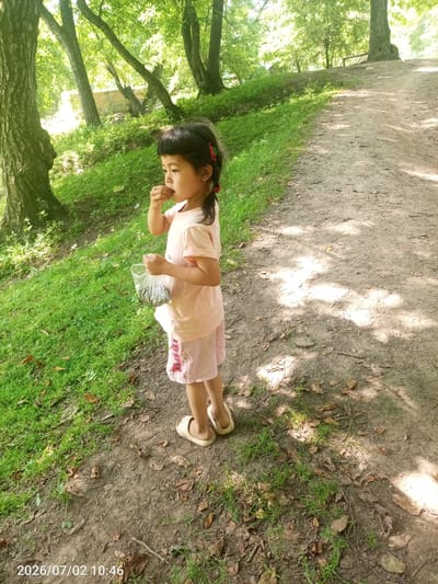 A young East Asian girl with dark hair styled in pigtails with red ties stands on a dirt path in a wooded area. She is wearing a light pink t-shirt and light pink shorts with a pattern. In her right hand, she holds a clear plastic bag filled with small, silvery objects, possibly fish. Her left hand is raised to her mouth, and she appears to be eating something. She is wearing light-colored sandals. The background consists of green grass, trees with dappled sunlight filtering through their leaves, and a dirt path. The timestamp "2026/07/02 10:46" is visible in the lower-left corner of the image. The location is identified as Жай-Терек, Kyrgyzstan.