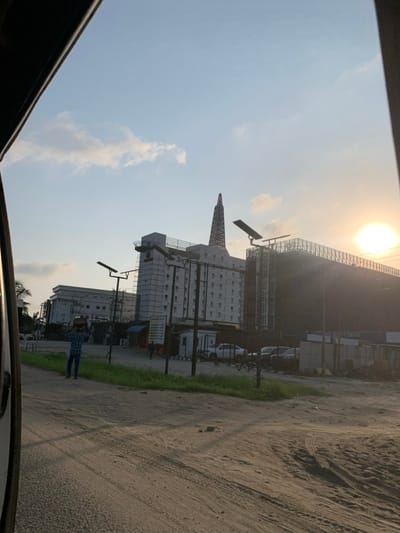 The image depicts an urban or semi-urban street scene in Ikota, Nigeria, under a pale blue sky with scattered clouds and a bright, low sun visible on the far right, suggesting late afternoon or early evening. The foreground features an unpaved, sandy-dirt road exhibiting tire tracks and a strip of green grass on the left.
In the mid-ground, a lone male figure walks leftward across the dirt road, carrying a dark, round object balanced on his head. He wears a plaid shirt and dark trousers. Further back and to his right, another less distinct individual is visible near a white, shed-like structure.
The background is dominated by several multi-story buildings. To the left-center, a light-colored building, possibly white or light grey, is partially covered in scaffolding, indicating construction or renovation. A distinctive, dark, spire-like structure, reminiscent of an oil derrick, rises from its roof. To the immediate right of this building, a large, dark-colored structure is heavily covered with dark netting and scaffolding, signifying extensive construction. A sign with "BEAUTY CENTER" is partially visible on this structure's lower facade. Several white sedans and other vehicles are parked in an unpaved lot in front of this large dark structure.
Multiple tall poles with solar panel fixtures are positioned throughout the scene, presumably for lighting. Patches of sparse green vegetation are present near the buildings and along the road. The extreme upper left and right corners of the image show parts of a dark, curved frame, likely from inside a vehicle, indicating the photograph was taken from a moving or parked car.