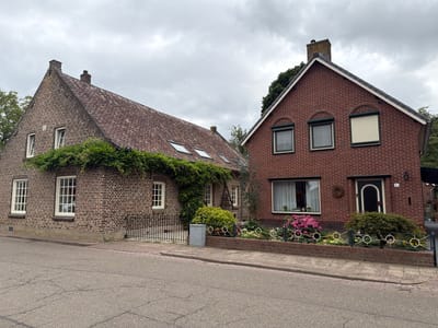 The image shows a street scene in Melick, Netherlands, with two attached brick houses on the right side of the road. The house on the left has a red tiled roof and is covered in ivy. It has white-framed windows. The house on the right is also made of red brick and has a pitched roof. It features several windows, some with rolling shutters. A small garden with pink flowers and green bushes is located in front of the right house, bordered by a decorative metal fence. The street in the foreground is asphalt with some cracks. The sky is overcast. There are no people visible in the image.