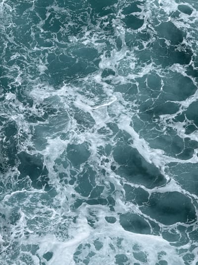 The media file is an abstract aerial shot of the ocean's surface, showcasing the intricate patterns of waves and foam. The dominant colors are shades of deep teal and turquoise from the water, contrasted with bright white from the churning foam and spray. The texture is varied, with smooth, dark patches of water interspersed with frothy, irregular shapes of white foam that create a dynamic, almost lace-like appearance across the frame.

The activity depicted is the natural movement of the sea, with waves creating eddies and currents that churn the surface into foam. There are no discernible subjects like people or objects. The setting is the open ocean.

Notable details include the abstract nature of the composition, which focuses on the organic and ever-changing forms of the water. The lighting suggests it could be daytime, possibly with some cloud cover or at an angle that diffuses direct sunlight, as there are no harsh shadows. The location is suggested as Llanes, Spain, which implies a coastal environment, likely the Cantabrian Sea. The image evokes a sense of powerful, natural forces and the beauty found in their chaotic yet organized patterns. There is no visible text in the media file.