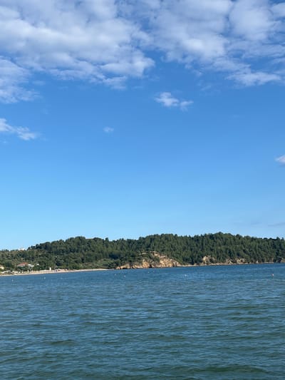 The image shows a wide view of the sea in front of a green, tree-covered hill. The sea is a deep blue with small ripples on the surface. In the distance, a sandy beach is visible, lined with white umbrellas and some buildings. There are a few small orange buoys in the water. The sky is clear blue with scattered white clouds. The location is the coast of Potos, Greece, with the Thassos island visible in the background.