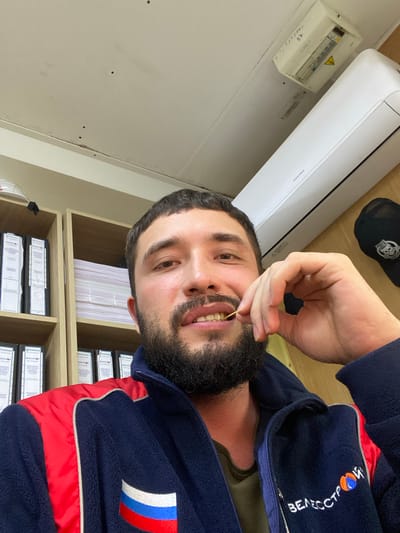 In Luzhitsy, Russia, a man with a beard is taking a selfie in an indoor office environment. He is wearing a dark blue fleece jacket with red accents and a Russian flag patch. He is holding a toothpick in his mouth. Behind him, there are shelves filled with binders and papers, an air conditioning unit mounted on the wall, and an electrical panel. A black baseball cap is visible on a wooden surface to the right.