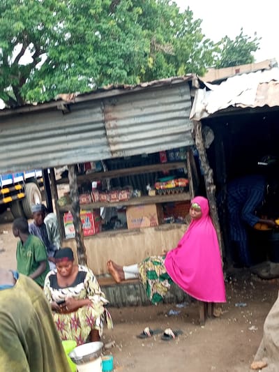 In Gadao, Nigeria, a street market scene unfolds under a corrugated metal roof supported by wooden poles. Several individuals are present. A woman wearing a bright pink hijab and a patterned dress is seated on a bench, leaning against a wooden post. To her left, a woman in a black, white, and yellow patterned dress is seated, looking at her phone. In the background, two men in green and grey shirts are standing, with a truck visible behind them. A shelf behind the seated women displays various items, possibly food products in colorful packaging. The ground is dirt, with some scattered debris.