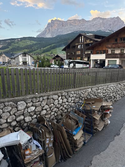 The image shows a street scene in the village of La Ila - Stern - La Villa, Italy. In the foreground, several metal trolleys are loaded with cardboard boxes and other recyclable materials, some of which are damaged or crushed. A stone wall with a wooden fence separates the street from a residential area. Behind the fence, there are several buildings, including a hotel with the sign "LA MAJUN" and a large mountain range in the background. The sky is partly cloudy. A bus and a few cars are visible on the street.