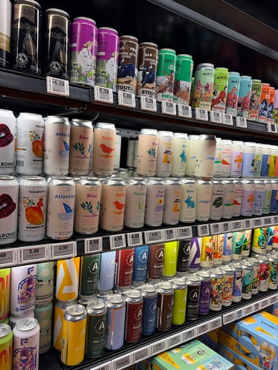 The image shows shelves in a refrigerated display case, likely within a retail store in Saint-Bruno-de-Montarville, Canada, filled with various brands and types of canned alcoholic beverages, primarily beer. The cans are arranged in rows, and each has a price tag attached to the shelf directly in front of it. Several brands are visible, including "ALA", "Boldwin", "Alchimist", and "Roquet". The types of beer vary, with labels like "IPA", "Stout", "Gose", and other descriptions indicating different flavors and brewing styles. The price tags show prices such as $3.99, $4.49, $4.99, $5.39, $5.49, $5.89 and $6.39. The lighting is bright, illuminating the cans and labels, creating a clear display for shoppers.