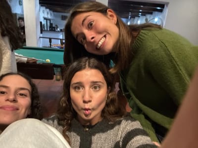 Four young women are arranged around the camera, their faces close together. One on the left wears a green sweater and smiles widely. Above her, another woman pokes out her tongue. To the right, a third woman smiles gently. A fourth woman is partially visible in the upper right corner, also smiling. A pool table with a yellow ball is visible in the background. The image is taken indoors, with stairs and decorations visible behind the subjects.