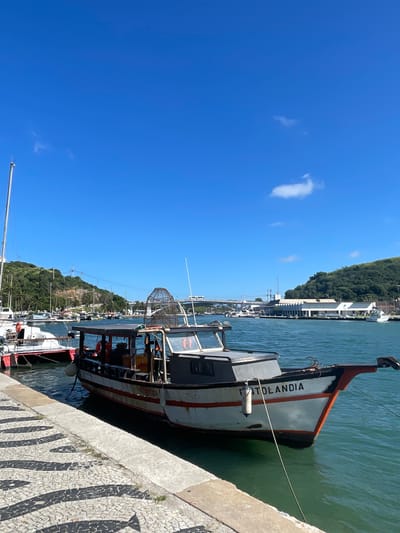 The image shows a boat named "FITOLANDIA" docked at a pier in Cabo Frio, Brazil. The boat has a distinctive orange and white bow. The pier's surface features a patterned black and white pavement. The water is green, and there is a view of green hills and buildings along the coastline. The sky is clear blue with few clouds. Other boats are visible in the background. People are visible inside the boat.