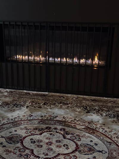 An interior scene is depicted within a structure in Maldegem, Belgium. A contemporary linear gas fireplace is centrally positioned in the upper half of the frame. The fireplace exhibits multiple visible gas flames emanating from a bed of white, granular, decorative media. A dark-colored, multi-paneled metal grate, composed of vertical bars, is situated directly in front of the fireplace, functioning as a protective barrier. The fireplace unit and the wall above it are dark-toned. The foreground consists of a patterned area rug with a light-colored, likely cream or beige, field. The rug features intricate floral and curvilinear motifs in shades of maroon, red, dark blue, and gold. A large circular or oval medallion design is partially visible on the rug in the lower-left quadrant. No human or animal subjects are present within the captured image. The ambient lighting is subdued, with the fireplace flames providing the primary illumination.