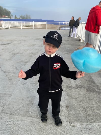 A young boy wearing a black baseball cap with a Mercedes-Benz logo, a black tracksuit with white trim, and black shoes stands on a concrete surface, holding a large piece of blue cotton candy. His eyes are closed, and he is giving a thumbs-up with his left hand. In the background, a white railing separates the walkway from a blue barrier, beyond which are trees and buildings. Several adults and a dog are visible in the distance. The sky is overcast. The scene appears to be outdoors, possibly near a waterfront, given the railing and the misty atmosphere suggesting a body of water, consistent with the location of Listvyanka, Russia, which is situated on Lake Baikal.