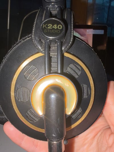 A close-up shows one earcup of an AKG K240 Studio headphone. The earcup is circular and primarily black, with a thin gold ring around the center. A cream-colored, crescent-shaped element is in the middle. The model number "K240 STUDIO" is visible on the black plastic connecting piece. A human hand is visible, partially covering the right side of the earcup. The location is unknown, as no information is provided about the specific location in L'Hospitalet De Llobregat, Spain or any other location.