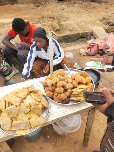 In Bokkos, Nigeria, a street vendor displays an assortment of fried snacks. Two individuals are visible in the foreground: a man in a red shirt and a woman wearing a black headscarf and a white and blue jacket. They appear to be engaged in a transaction with a customer whose hand is holding money and a smartphone.
The snacks are arranged on two metal platters on a weathered wooden table. The platter on the left contains slices of what appears to be fried plantain or yam. The platter on the right holds a mixture of round, golden-brown fried items and some flatter, fried pieces. The background shows a dirt ground with some debris and a pink tarp. The scene suggests a bustling marketplace or roadside stall, typical of urban or semi-urban Nigerian settings.