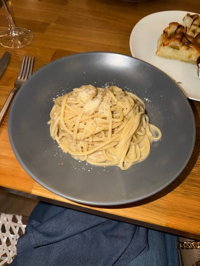 The image displays a dining scene at an Italian restaurant in Las Vegas, United States. In the foreground, a dark grey circular plate holds a serving of spaghetti coated in a light-colored, creamy sauce, liberally topped with grated cheese and flecks of black pepper. To the right of the main plate, a slice of focaccia bread, garnished with olives and herbs, rests on a white circular plate. A silver dinner fork and knife are positioned to the left of the spaghetti plate, lying on a light-toned wooden table surface. A clear, stemmed wine glass is visible in the upper left quadrant. Partially visible below the table is the lap of an individual, dressed in dark blue denim fabric, with a section of a light-colored woven chair base adjacent. A dark grey handbag with a gold-colored chain strap is partially visible on the right side of the person's lap.
