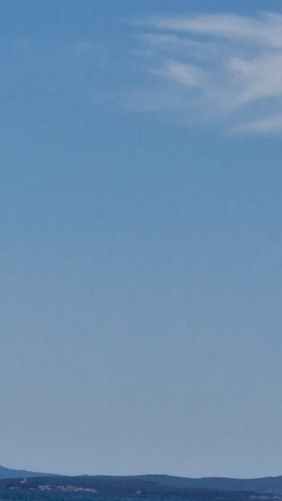 The image displays a broad view of the sky above the Adriatic Sea, with a distant coastline of Zadar, Croatia, visible at the bottom. The sky is a pale, clear blue, with scattered, wispy white clouds concentrated in the upper right quadrant. The sea is a dark, rippling blue, meeting the landmass which appears as a dark, undulating silhouette. On the land, faint details of buildings and structures can be discerned along the coast, suggesting a populated area. The overall scene is serene and expansive, conveying a sense of natural beauty and distance.