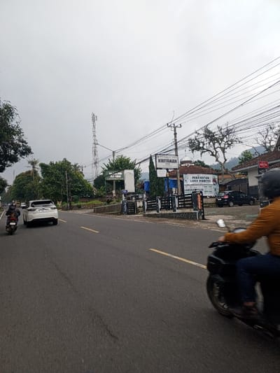 The image depicts a street scene, identified as being in the city of Qorday, Kazakhstan, though prominent signage in the Indonesian language suggests a different geographical context. The environment is an overcast daytime setting with a grey sky.
In the foreground, a two-lane asphalt road with a solid yellow center line extends into the distance. On the right side of the road, a person wearing a brown jacket and dark helmet is riding a black scooter, partially visible and blurred due to motion, traveling from right to left. Further down the left lane, a white SUV with a visible license plate (starting with "B") is traveling away from the viewer. Behind the SUV, another individual on a motorcycle is also moving away.
The right side of the road features a paved shoulder leading to a series of buildings and structures. A utility pole with numerous overhead power lines stands prominently. Several signs are visible: a white sign with black text reads "KHITANAN," and another large blue and white sign displays "PERAWATAN LUKA DIABETES" and "LABORATORIUM." A sign for "MASJID AL-ILM" is also present. Behind these signs, buildings with light-colored walls and red-tiled roofs are visible, along with what appears to be a mosque's dome. A black car is parked further down the right side.
The left side of the road is bordered by dense green foliage and trees, indicating a lush landscape. In the distant background, a tall, grey cellular or communication tower stands against the sky, with hills and more greenery visible behind it.