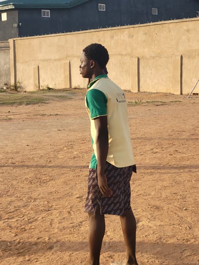 A young Black man stands on a dirt ground in Ibo, Nigeria. The man wears a two-toned shirt with a light yellow body and green sleeves, and dark shorts covered in the Louis Vuitton logo. He is facing the left of the frame, standing perpendicular to a beige building with regular wall indentations. Behind the building and to its left is a building with a dark-colored siding. Several window panes are visible on this building. The ground is dry and cracked, and there is a small patch of grass between the building and the ground.
