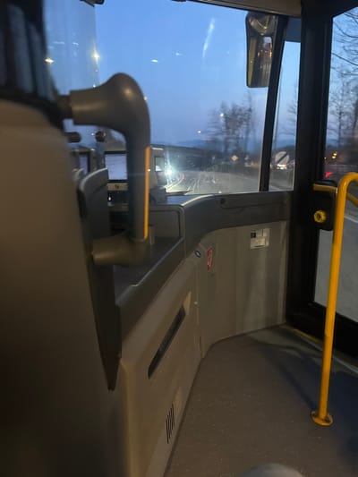 The image provides a forensic view from inside a public transport vehicle, likely a bus, operating in Krems an der Donau, Austria. The perspective is from a passenger seat positioned behind the driver's area, looking forward and slightly right, through the front windshield and a side window.
In the immediate foreground on the left, the upper back of a light-colored seat is visible. Extending from the mid-left is a grey vertical pole, possibly a grab handle or seat support, featuring a distinct yellowish reflective strip. To the right of this pole, the driver's console is partially visible, displaying a digital screen with white text against a dark background, and an adjacent ticketing or card reader device.
The right side of the interior features a closed door. A bright yellow vertical grab pole is securely mounted to the dark grey, speckled floor and extends upwards, with a yellow circular mechanism near its base. Light grey wall panels display red-bordered emergency pictograms, including a fire extinguisher symbol and another generic emergency sign, along with a small white rectangular label. A horizontal vent grille is present on the lower part of the left wall panel.
The exterior view, observed through the vehicle's windows, indicates twilight or early morning conditions, with a pale blue sky dominating the upper background. An asphalt road is visible ahead. Bare trees and vegetation line the right side of the road. In the distance, an overpass or bridge structure traverses the scene. The distinct headlights of at least one oncoming vehicle are visible beneath the bridge. Traffic signage, including a yellow diamond-shaped warning sign and a circular red-bordered prohibitory sign, are partially discernible. Distant streetlights or other artificial light sources appear as bright points. The front windshield exhibits minor smudges or condensation trails. No individuals are explicitly visible.