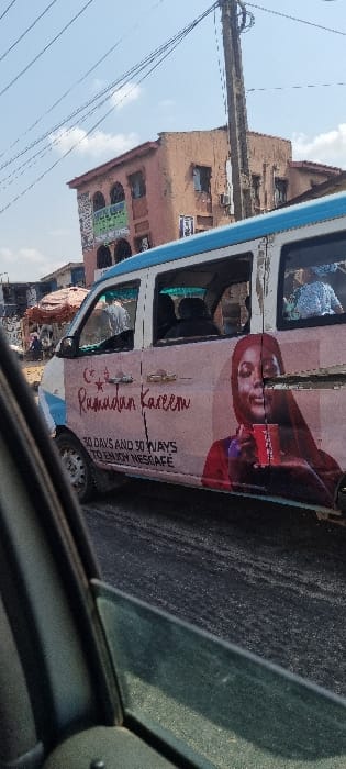 This image presents a daytime street scene in Ojodu, Nigeria, observed from inside a vehicle. In the midground, a white and light blue commercial passenger van is visible on a dark asphalt road. The van's side is extensively covered by an advertisement featuring a woman in a red hijab holding a red cup, accompanied by text reading "Ramadan Kareem" with a crescent moon and star symbol, and "30 DAYS AND 30 WAYS TO ENJOY NESCAFÉ." Multiple passengers are partially visible through the van's windows. To the immediate left background, a busy street market area with pedestrians and stalls, including a brown tent-like structure, is discernible. To the right background, a two-story building with a light brown facade and blue security grilles on its windows stands prominently, with a green and white commercial sign affixed. A tall utility pole with numerous overhead wires dominates the space between the van and the building. The sky above is bright blue with scattered white clouds.