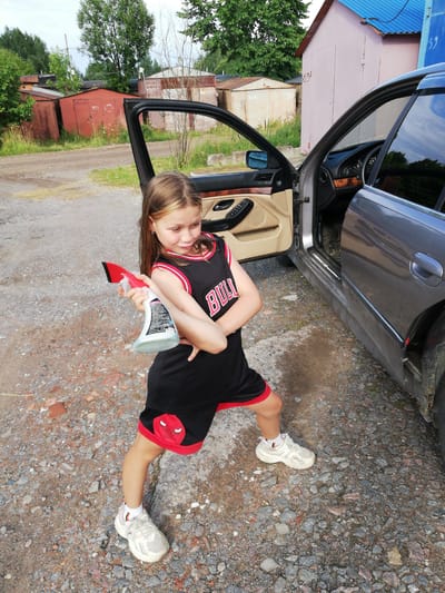 A young girl with blonde hair and a black and red basketball jersey stands next to the open door of a dark gray car. She is holding a red and white spray bottle in her right hand and her left arm is bent with her hand on her hip. The girl is wearing white sneakers. The scene appears to be in a rural area with gravel ground and several garages in the background. The sky is overcast. The girl's jersey has the word "BULLS" across the chest. The shorts have red trim and a red bull logo on the side. This location is identified as the city of Tosno, Russia.