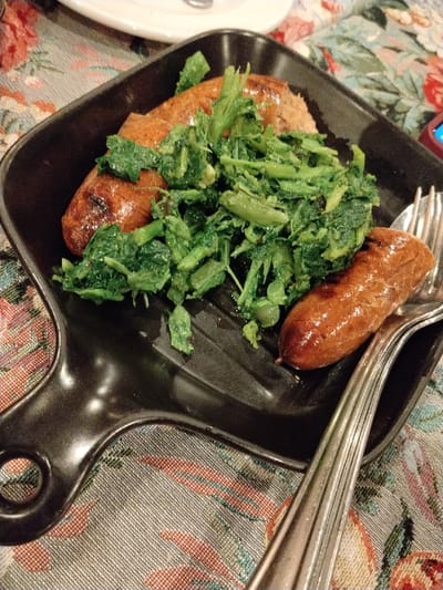 A black, cast-iron skillet holds a meal of grilled sausages and leafy green vegetables, possibly broccoli rabe. Three sausages are visible, browned with grill marks. The greens are piled in the center, appearing sautéed. A silver fork rests on the skillet, touching the sausage on the right. The skillet sits on a floral patterned tablecloth, and a white plate is partially visible in the background.