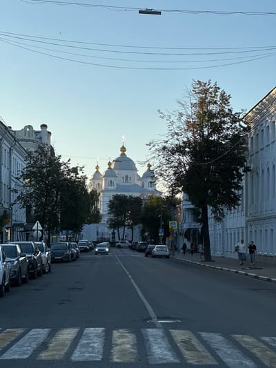 The image shows a street scene in Yazvitsevo, Russia, with a white domed church visible in the background. The church has several golden domes and spires, and is surrounded by trees. On the right side of the street, there are white buildings with arched windows. On the left side of the street, there are parked cars. The street has a pedestrian crossing in the foreground. Several people are walking on the sidewalk on the right side of the street. The sky is clear and blue, and the sun is shining. The scene appears to be set during the daytime, possibly in the late afternoon or early evening due to the lighting.